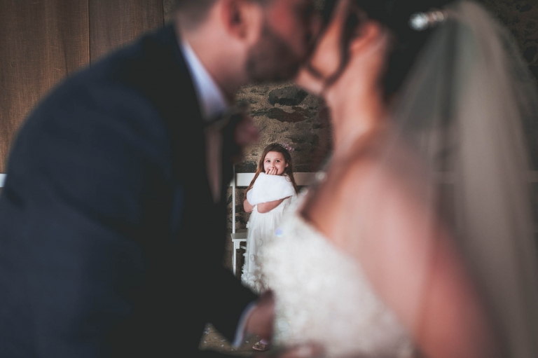 Flower girl giggles as she looks at bride and groom kissing
